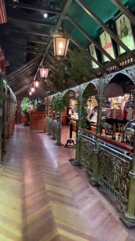 The video showcases an elegant and ornate indoor buffet area, likely within a high-end restaurant or hotel. The setting is characterized by a luxurious, traditional design with a greenhouse-like structure overhead, featuring a green canopy and decorative metalwork that resembles intricate wrought iron or bronze. Hanging lanterns with warm light illuminate the space, and some greenery, possibly vines, are intertwined with the overhead structure.

The flooring is made of polished wood laid in a herringbone pattern. Along the right side of the video, a long buffet line is set up. This line features various food stations with shiny silver chafing dishes and decorative copper pots, suggesting a variety of hot food options. Ornate brass or bronze railings, topped with wooden handrails, separate the main walkway from the buffet area. Red velvet ropes are visible, indicating queuing areas or a barrier to prevent direct access to the food until served or to guide traffic flow.

Several staff members are present. One individual, dressed in a white shirt and black apron, is actively working at a food station, appearing to arrange or prepare items. In the background, another staff member in a white chef's coat and hat can be seen. The buffet line also includes a section with smaller prepared items, possibly desserts or appetizers, arranged neatly on trays. The overall ambiance is sophisticated and inviting, with warm lighting and rich details in the decor.