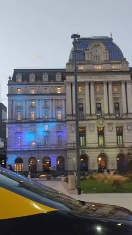 The video presents an evening view of the Secretaría de Comunicaciones building in Buenos Aires, Argentina. The imposing structure is illuminated with lights, with blue hues highlighting the left side of the building. The building features a distinctive dome with a clock and the inscription "Secretaría de Comunicaciones."

In front of the building is a statue of Juana Azurduy on a base, surrounded by grass and tall vegetation. People are seen walking and sitting around the area, enjoying the evening ambiance.

A black and yellow taxi is parked in the foreground on the left, providing a sense of urban activity. The sky is overcast, indicating that it is dusk.