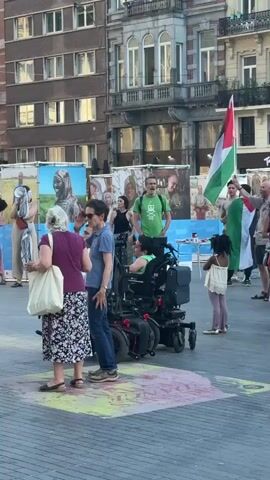The video shows an outdoor gathering in a city square. In the foreground, several people are standing, some holding Palestinian flags. One person is in a wheelchair. In the background, there are large banners and ornate buildings with classical architecture. The banners display text in what appears to be multiple languages. One banner reads "PROOST! SANTE! CHEERS!" followed by "Belgian Beer World Experience." The other banner appears to advertise "150 BELGIAN BEERS." 

The scene suggests a public event or demonstration, possibly related to a cultural or political cause, given the prominent display of Palestinian flags. The presence of a wheelchair user and other individuals suggests a diverse group of participants. The time of day appears to be daytime, with sunlight casting shadows. The weather seems pleasant, suitable for an outdoor gathering. The architectural style of the buildings and the banners suggest a European setting, consistent with the provided location context of Brussels, Belgium. There is a mix of individuals of different ages and genders, and their expressions are largely neutral or engaged in the activity.