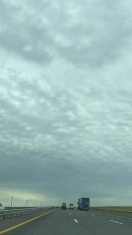 The video shows a view from a vehicle driving on a highway under a cloudy sky. The highway has multiple lanes, marked with yellow and white lines. A metal guardrail runs along the left side of the road. Several vehicles are visible on the highway, including a blue truck with a trailer, a white van, and other cars and trucks in the distance. The sky is filled with scattered clouds, giving a somewhat overcast appearance to the scene. The landscape on either side of the highway appears to be flat and open, with some vegetation. The camera perspective suggests the video was recorded from the driver's seat or a passenger seat of the moving vehicle.