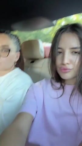 The video shows three young women in the back seat of a car. The woman in the foreground on the right has long, dark hair and is wearing a pink t-shirt. She is looking at the camera and smiling slightly, with her lips slightly pursed. The woman next to her, on the left, has her hair pulled back in a ponytail and is wearing glasses and a white sweatshirt with red writing. She is looking to the left and her mouth is slightly open as if she is speaking or singing. The third woman is partially visible behind the woman with the ponytail, with blonde hair, and appears to be asleep or resting her head. The interior of the car is visible, including leather headrests and the window, through which trees and greenery can be seen. The lighting suggests it is daytime. The woman in the pink shirt waves her hand.