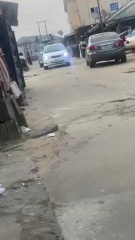 The video depicts a narrow, rundown street in what appears to be a developing nation. The road surface is cracked and uneven, with scattered litter. Buildings of varying conditions line the street, including a multi-story building with a red roof. Several cars are parked along the street, and one white car is driving towards the camera. A woman in a long dress stands near a makeshift structure on the left side of the frame, and a man wearing blue jeans is partially visible beside her. The sky is overcast.