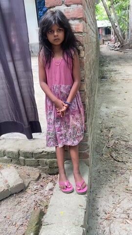 A young girl stands in a yard. She is wearing a pink floral dress, a gold necklace, and a blue bracelet. She also has on pink flip-flops. Her dark hair is down around her shoulders. She is standing on a concrete block next to a brick wall. To her left is a dark curtain. Behind her is a dirt path lined with trees. She looks off to the right, then looks to the camera and opens her mouth, like she's speaking.
