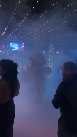 A mascot costume character, possibly a cartoon character, stands in the center of a room filled with fog and blue and purple lighting. The character is wearing a light-colored outfit and appears to be interacting with the environment. String lights are visible across the ceiling. In the background, there are screens displaying images, stage lighting equipment, and people dancing or standing around. Some individuals are holding drinks and fans. The overall atmosphere suggests a party or event with entertainment.