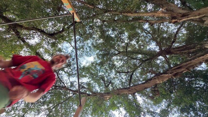 A low-angle shot looks up into a canopy of tree branches, revealing a person hanging from a metal pull-up bar. The person is wearing a red shirt and green shorts. He jumps and flips 360 degrees around the bar, facing the camera as he completes the rotation.