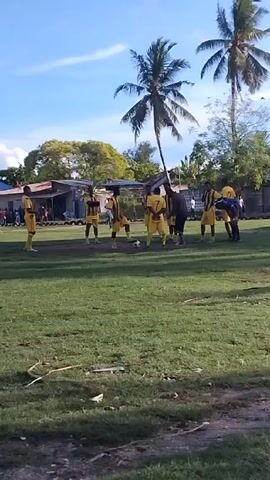 The video captures a soccer game taking place on a grassy field. A group of soccer players, dressed in yellow and black striped jerseys, are gathered around a soccer ball. The field is surrounded by spectators, and buildings and trees are visible in the background. The sky is blue with scattered clouds.