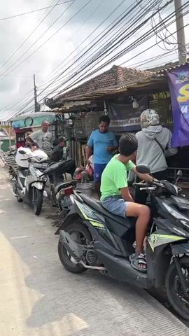 A group of people are gathered on a street in front of a building that appears to be a shop. Several motorcycles are parked in a line. Some individuals are standing, while others are sitting or on their motorcycles. One person is wearing a bright green shirt and is sitting on a black motorcycle. Another person in a grey hoodie is also near a motorcycle. The building has a tiled roof and a sign that is partially visible. Overhead, there are many electrical wires. The overall impression is a busy street scene with people interacting and vehicles present.