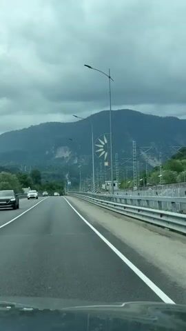 The video shows a car driving on a highway on a cloudy day. There are several cars on the road, and a mountain range can be seen in the background. On the right side of the road, there are train tracks with overhead electrical wires. The car continues down the road.