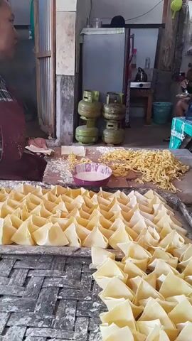 The video shows a domestic kitchen or food preparation area. In the foreground, there are rows of folded, uncooked wonton wrappers or dumplings arranged on a metal tray dusted with flour. Behind the tray, a bowl of white liquid, possibly egg wash or a binding agent, is visible, along with a pile of cooked noodles or dough. In the background, a person stands by a refrigerator, and further back, two men are seated at a table. One of the men is looking at a tablet or phone, while the other is also seated. There are several green gas canisters stacked against a wall. The environment appears to be rustic, with exposed wooden beams and concrete walls. The overall scene suggests a food production or preparation process.