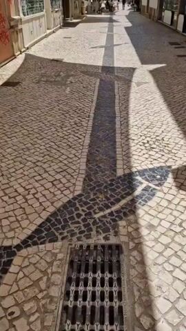 The video shows a POV shot of walking down a narrow cobblestone street. The street is lined with buildings and has a black and white mosaic pattern running down the center, forming a path. Shadows from buildings and possibly awnings are cast across the pavement. In the distance, there are a few people walking and sitting at tables outside. The sun is bright and casts sharp shadows. There is a drain grate visible on the ground in the foreground.
