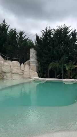 A video transitions between two scenes. The first shows a tropical pool area with palm trees, thatched umbrellas, and lounge chairs. The second scene shows a wider view of the same area, revealing a small swimming pool, more lounge chairs, and a panoramic view of a town nestled in rolling hills under a cloudy sky. Lush greenery and palm fronds frame the view.