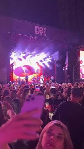 The video shows a concert. A band is performing on a stage illuminated by bright red and blue lights. The band consists of multiple members, including singers and instrumentalists. In front of the stage, a large crowd of people are watching the performance. Many of them are holding up their cell phones, presumably to record or take pictures. The crowd appears to be enjoying the show, with some people dancing and cheering. The overall atmosphere is energetic and lively.