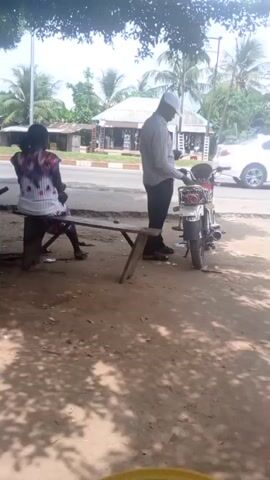 The video shows a man in a light-colored shirt and white cap standing next to a small motorcycle on the side of a road. A woman is seated on a wooden bench nearby. The area appears to be a roadside shaded by trees, with other buildings and vegetation visible across the road. A three-wheeled vehicle drives by on the road. There's a sign propped up that reads "POSI" with services listed below. The ground is dirt and covered with shadows from the trees above.
