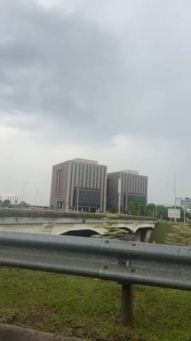 The video shows a view from behind a metal guardrail, looking across a bridge towards two square office buildings with beige and grey exteriors. The bridge is adorned with ornate white decorations and green vegetation. The sky overhead is overcast with grey clouds. Streetlights are visible along the roadway near the buildings. A vehicle is briefly seen crossing the bridge. The scene suggests a modern urban landscape under gloomy weather conditions.