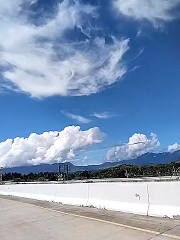 The video shows a view from a car traveling on a highway. The sky is blue with fluffy white clouds. In the distance, there are mountains and trees. A road sign is visible above the highway. The road is smooth and gray with white dividers. A low white wall lines the side of the highway. The video is taken from a low angle, emphasizing the sky and the surrounding landscape.