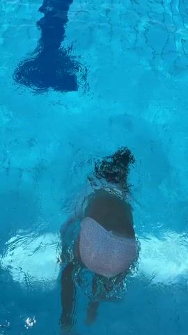 The video shows a person swimming underwater in a blue swimming pool. The person is wearing a swimsuit and appears to be performing a somersault or similar maneuver, with their legs kicking upwards at one point and then their arms extending outwards. The water is clear, and the bottom of the pool is visible. Sunlight creates patterns of light and shadow on the water's surface.