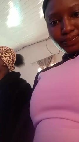 The video shows two people in what appears to be an indoor setting. The person in the foreground is wearing a pink outfit and large hoop earrings. Their hair is covered with a black head covering. Beside them and slightly behind is a person wearing a black jacket over a patterned top and a headscarf with a brown and white design. They are both looking forward, suggesting they are watching something. The background is plain, with a white ceiling with grooved panels.