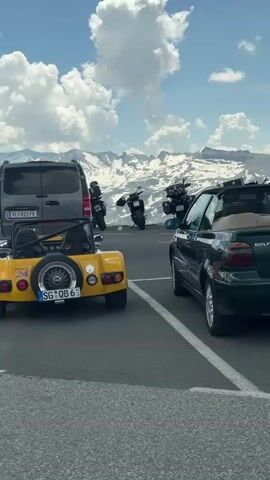 The video shows a parking area on a mountaintop with a clear blue sky and fluffy white clouds. Snow-capped mountains are visible in the background. Several vehicles are parked, including a yellow three-wheeled car, a dark gray van, a dark green convertible, and a dark gray sedan. A group of people, some wearing helmets and motorcycle gear, are gathered around the vehicles, appearing to be taking a break or admiring the view. The yellow car has a spare tire mounted on the back and a license plate from Switzerland. The dark green convertible's top is down. A person in a bright yellow jacket is standing by the dark gray sedan, and another person is standing by the dark gray van. A few motorcycles are also parked in the area. The overall scene suggests a scenic stop during a road trip or motorcycle tour.