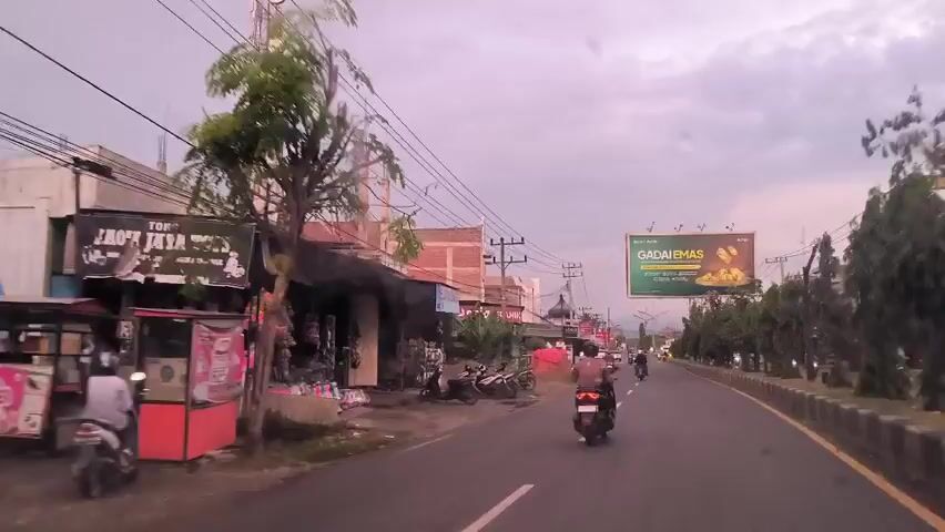 The video shows a view from inside a vehicle driving down a street. The street is lined with buildings and shops on one side and a grassy curb with trees on the other. A large billboard advertising "GADAIEMAS" is visible on the right. Several motorcycles are driving along the street, both ahead of and behind the vehicle with the camera. The sky is overcast and the light is subdued, suggesting it might be early morning or late afternoon. The road has clear white lane markings. Shops on the left side of the road are visible, some displaying goods for sale. Overhead power lines run across the street.