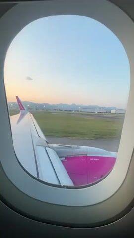 The video shows a view from an airplane window. The airplane is on the ground, likely taxiing or waiting to depart, as indicated by the runway visible outside the window. A portion of the airplane's wing, painted white with a pink section, is visible in the foreground. The pink section has the text "NO STEP" printed on it. Beyond the wing, a grassy area and a tarmac with runway markings can be seen. In the distance, there are buildings, possibly airport infrastructure, and a range of hills or mountains against a pale blue sky with hints of pink and orange from a sunrise or sunset. The light suggests it is either early morning or late evening.