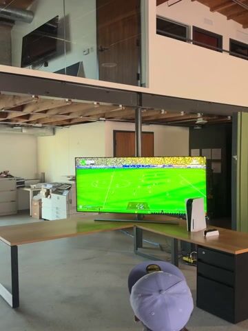 The video shows the interior of a large open office space. A large TV with a soccer game playing is visible near a desk with a Playstation 5 console. The camera pans to show a snack bar area with shelves, coolers, and a person in a yellow shirt arranging items. The space has high ceilings with exposed wooden beams and various lighting fixtures. Walls are painted in neutral colors.