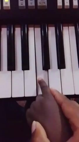 The video shows a close-up of a person’s hands (presumably an adult) guiding the hand of a child over the keys of an electronic keyboard or organ. The keyboard is black and white, and has lighted buttons above the keys, numbered 1 through 6. The person’s hand is dark-skinned, and the child’s hand is also dark-skinned.  The hands press the white keys in succession, from left to right.  The background is dark and out of focus.