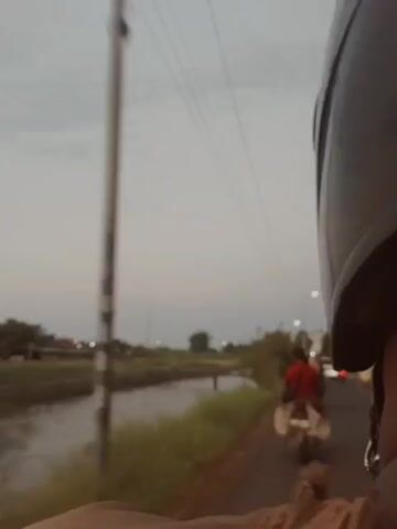 A first-person perspective video shows a person riding on a motorcycle. The rider is wearing a red shirt and is seated behind another individual. The motorcycle is traveling on a paved road alongside a body of water, likely a canal, which is bordered by tall grass. In the distance, there are buildings and trees. Power lines are visible overhead against a cloudy sky. The camera is mounted on the motorcycle, facing forward and slightly to the right, capturing the passing scenery. The video is recorded at dusk or in overcast conditions, with the lighting being dim.