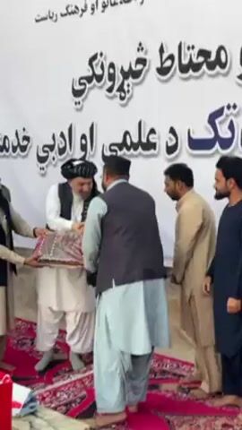 The video depicts an awards ceremony or a similar event taking place in Kandahar, Afghanistan. The setting appears to be indoors, with a large white banner in the background featuring text in Pashto. The banner's text translates to "Ministry of Information and Culture, Republic of Afghanistan" and "Official News Agency" at the top, followed by "Professional Cultural and Literary Services" and "Cultural and Literary Services" in larger font.

In the foreground, several men are gathered. The central action involves an older man, dressed in traditional Pashtun attire with a white shalwar kameez and a dark turban, receiving an item wrapped in patterned cloth from another man. Both men are smiling, suggesting a positive and respectful interaction. The recipient of the award is also wearing a dark vest over his traditional clothing.

To the left, another man, also in traditional attire, appears to be presenting the award. On the right, two other men are observing the ceremony. They are also dressed in traditional clothing, with one wearing a light-colored shalwar kameez and the other a dark blue one. The ground is covered with a red rug with intricate patterns. A red gift bag with a "G" logo is visible at the bottom left. The lighting suggests it is daytime, and the atmosphere seems formal yet warm, indicative of a significant cultural or official event.