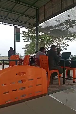 The video shows an outdoor seating area with several orange plastic chairs and tables. In the background, there are people sitting at tables. Some individuals are using laptops. A person wearing a hijab is standing near a table. The area appears to be covered by a roof. The sky is overcast, and there are trees and foliage visible in the distance. The overall scene suggests a casual gathering or a place for people to work outdoors.