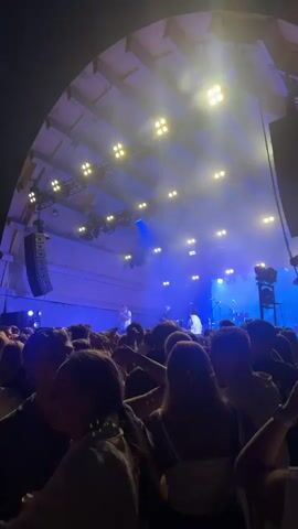 This is a shaky, handheld video shot from within a crowd of people attending an outdoor concert at night. The stage is visible in the distance with bright blue and white lights illuminating it. Several performers are on stage, but they are indistinct due to the distance and blurriness of the video. The crowd is dense, with many people facing the stage. The atmosphere appears to be energetic and celebratory. The background consists of trees and a dark sky with some string lights visible. The video is mostly composed of close-ups of the backs of people's heads and shoulders, with occasional glimpses of the stage and surrounding environment.