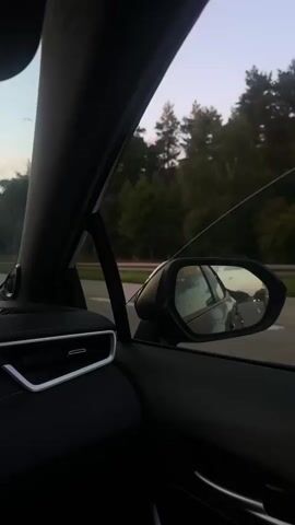 The video is a first-person perspective shot from inside a car. The car is moving along a highway. On the left side of the frame, the dashboard and part of the driver's side door are visible, including an air vent with chrome trim. The car's side mirror is visible on the right, reflecting the road and traffic behind. Outside the car window, a dense forest lines the highway, and the sky is a soft, dusky orange and blue, suggesting sunset or sunrise. The overall scene conveys a sense of travel and transition.