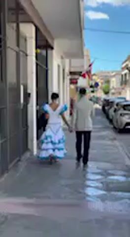 In a daytime video clip filmed in Jerez de la Frontera, Spain, two individuals walk away from the camera down a sunny paved sidewalk. The person on the left is dressed in a vibrant, traditional Spanish-style dress, featuring a white bodice and a full, multi-tiered skirt with white and blue ruffles. They appear to have a dark bag slung over their shoulder. The person on the right wears a light-colored jacket or shirt with dark trousers. Both are walking close together, possibly holding hands, along a building with dark-framed windows or doors. To their right, a street with parked cars is visible under a clear blue sky with some scattered clouds. The scene suggests a pleasant day in an urban or town setting, with one individual in attire typical of a festive or cultural event in the region.