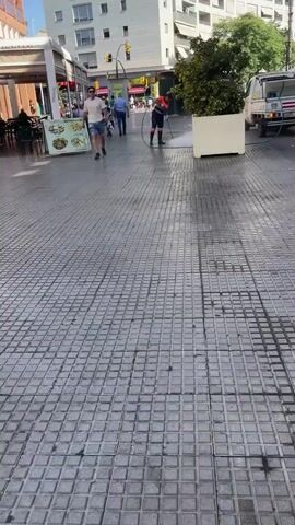 A person in a safety vest and dark pants is pressure washing a tiled pedestrian area. The person is holding a cleaning wand and directing a powerful stream of water onto the ground. In the background, a white truck with some equipment on its bed is parked. Several people are walking in the background. A man in a white t-shirt and denim shorts is walking towards the camera. The scene appears to be an outdoor public space, possibly a city street or plaza.