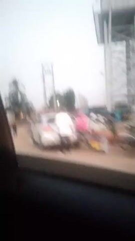 The video, shot from inside a moving vehicle, shows a street scene in what appears to be a town. There are people walking around, a silver car parked on the side of the road, and a motorcycle driving by. The buildings are low-rise and the sky is overcast. There is a low concrete wall with red and white painted blocks in the foreground.  The overall impression is of a street in a developing country. The image is somewhat blurry due to the motion of the vehicle.