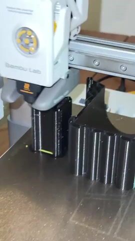The video shows a close-up view of a 3D printer, specifically a Bambu Lab brand, in operation. The printer's print head, equipped with a nozzle that emits molten plastic, is actively creating a black, ribbed object on a textured print bed. The object appears to be segmented or composed of multiple identical units arranged in a line. The print head moves smoothly across the surface, depositing filament layer by layer. The surrounding environment is out of focus but suggests an indoor setting, possibly a workspace or hobby area. The lighting is adequate, highlighting the details of the printing process.