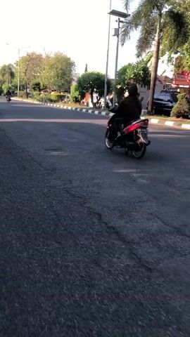 The media file shows a person riding a red scooter on a paved road. The rider is wearing dark clothing and a hijab. The scene appears to be a street in a suburban or semi-urban area, with trees, sidewalks, and buildings visible in the background. A street lamp is also visible. There is another person on a scooter further down the road, and a car parked on the side. The lighting suggests it is daytime, likely late afternoon or early morning, as the sun is casting long shadows and the sky is clear. The overall atmosphere seems calm and ordinary, depicting everyday life in a residential area. There is a sign with "Alfamart" written on it in the background, indicating a commercial establishment. The location is Klurak, Indonesia.