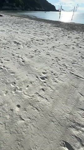 A first-person perspective video shows a hand scooping up sand on a beach. The sand is then let to fall through the fingers. In the background, a volleyball net with a sign hangs over the water. A hill covered in trees is visible on the left side of the frame, and the sun shines brightly in the sky.