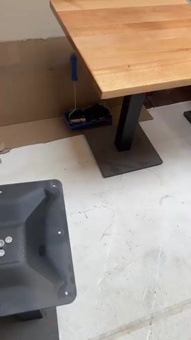 The video shows a collection of square wooden tables with black metal bases. The tables are arranged in rows, with some on the left side of the frame and more extending into the background. The wood appears to be a light-colored hardwood, with visible grain patterns. The tables have a smooth, finished surface.

The setting appears to be a workshop or storage area, with plain walls and a concrete floor that has some marks and smudges. Cardboard boxes are visible in the background on the left side. The lighting suggests an indoor environment, possibly during the daytime.

The activity taking place is the presentation or arrangement of these tables. The camera pans across the tables, showcasing their construction and how they are positioned. There are no people clearly visible in the frame, although there might be a person's feet or legs partially visible in the far background of the right side of the frame at the 00:04 timestamp.

Notable details include the consistent design of the tables, with a square wooden top and a central black pedestal base. The camera movement is smooth, giving a clear view of the objects. There is no visible text in the media file. The overall impression is one of newly manufactured or prepared furniture, likely for a restaurant, cafe, or event space. The location context of Waregem, Belgium is provided separately and is not discernible from the visual content itself.