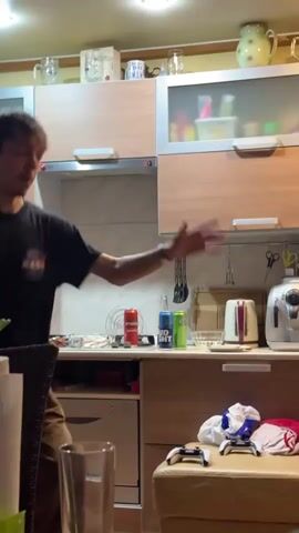 A young man is enthusiastically dancing in a home kitchen. He wears a black t-shirt with "NASA" printed on it and brown pants. He moves his arms and legs rhythmically, appearing happy and energetic.

The kitchen features light wooden cabinets with some frosted glass doors on the upper units. A white electric kettle, a silver toaster-like appliance, and two beverage cans (one labeled "BUD LIGHT") are visible on the counter. Kitchen utensils hang from a rack on the wall. In the foreground, a clear glass sits on a table, and to the right, two white PlayStation controllers and some folded clothes rest on what appears to be a couch or stool. The scene is illuminated by artificial lighting, suggesting it is evening or night.