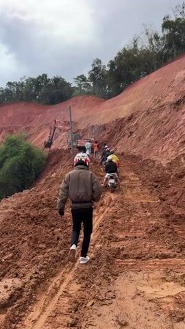A group of people are seen on a muddy, hilly road with a construction excavator in the background. One person is walking up the hill away from the camera, while several others are riding motorcycles up the same path. The terrain is steep and appears to be under construction or has experienced a recent landslide, with loose dirt and mud covering the road. The excavator is positioned on the side of the hill, appearing to be involved in earthmoving activities. The sky is overcast, and there are trees and vegetation on the higher slopes of the hill. In one shot, a motorcycle is seen overturned in the mud, suggesting difficult conditions.