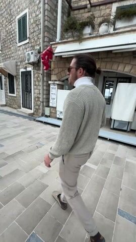 A man with long hair tied back in a bun, wearing glasses, a white turtleneck, a light-colored knitted cardigan, and light beige trousers, walks along a paved pedestrian area. He is followed by a small, fluffy, reddish-brown dog wearing a dark harness.

The scene is an outdoor setting, likely a town square or pedestrian street, characterized by stone buildings. On the left, a building features stone walls, a green shuttered window, and an air conditioning unit. A red object, possibly a decorative light or speaker, hangs from the building's facade.

On the right, there's a shop or cafe with arched entrances built into a stone structure. White protective covers or tarps obscure parts of the shopfront, possibly covering outdoor seating or equipment. Several signs are visible:
*   A vertical sign on the left side of the shop lists menu items such as "PANINI," "JUICE BAR," "BEER BAR," "ICE CREAM," and "SUNDAES."
*   Below it, another sign includes the words "CORNER AUTHENTIC."
*   A large, colorful ice cream cone decoration is mounted on the wall above one of the shop's arches.
*   Another menu board is partially visible on the right side of the shop.

The man and dog walk from the camera's perspective, then the man briefly stops and looks down at the dog. The dog also pauses, looking up at him. They then continue walking slowly past the shop, with the dog trotting beside the man. The paving stones are light-colored and laid in a distinct pattern. The overall lighting suggests it is daytime, possibly overcast.