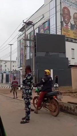 The video shows a street scene with a modern building covered in political posters. The building has multiple floors and is adorned with large banners featuring portraits of politicians and campaign slogans. The posters appear to be for the All Progressives Congress (APC) party, with names like "Bola Ahmed Tinubu As President" and "Governor, Ipinle Ogun" visible. Several flags are also displayed in front of the building. A man in a striped shirt and shorts walks down the street carrying a round object. The road in front of the building is paved but appears to have some potholes and debris. The sky is overcast. The video seems to be recorded from a moving vehicle, as indicated by the slight camera shake and the perspective.