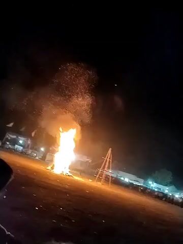 The video shows a nighttime scene of an outdoor event. In the foreground, the silhouette of people is visible, some of whom appear to be holding items. In the mid-ground, a large bonfire is burning brightly, casting an orange glow. Behind the fire, there is a structure that resembles a wooden frame or scaffolding. Further in the background, a line of people is visible, and what appears to be a stage or platform with lights is present. The ground is a dark, flat surface, possibly dirt or grass. The overall atmosphere suggests a festival or celebration.