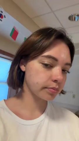 This is a short, selfie-style video of a young person with short, dark brown hair. They are wearing a white t-shirt with text and an image on it, and they have a light-colored tote bag slung over their left shoulder. The background appears to be an indoor setting, possibly a common room or rec area. The person looks at the camera, sticks out their tongue, and makes a playful face at different points in the video. In the background, there is a dartboard, a counter or desk, and some wooden paneling. The lighting is bright and appears to be natural or from overhead lights.