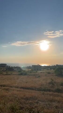 This is a vertical, medium shot video of a landscape. The sun is visible in the sky, casting a bright glow over the scene. There are some buildings in the distance, and the land is covered in dry grass. The video pans slightly to the left, revealing more of the landscape and the ocean.