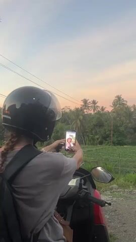 A person wearing a black helmet and a grey t-shirt, with a backpack on, is sitting on a motorcycle. They are holding a smartphone in their right hand, taking a selfie video. The person is facing away from the camera, looking at the phone screen. The background shows a rural landscape with green fields, palm trees, and a sky with a soft, pastel-colored sunset. The motorcycle is red and black. The video is brief, lasting only a few seconds.
