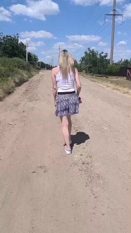This media file shows a woman walking away from the camera down a dirt road. She has long blonde hair and is wearing a white tank top and a black and white patterned skirt. The road is unpaved and dusty, with some sparse vegetation on either side. In the background, there are trees, power lines, and a blue sky with scattered clouds. The sun appears to be shining brightly, indicating it is daytime. The setting suggests a rural or semi-rural area. The woman is walking at a normal pace, and her posture does not convey any particular emotion, appearing to be a casual walk. There is no visible text in the media file. The approximate location is Varnița, Moldova, which is a village in the Transnistria region.