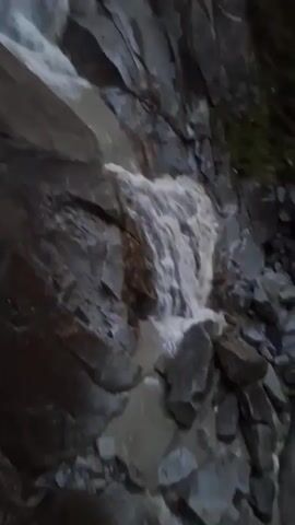 The video shows a rocky waterfall cascading down a mountainside. The water is a milky white, suggesting it may be carrying sediment. The rocks are dark gray and appear wet. There are some patches of green vegetation on the left side of the frame. In the distance, a small arched bridge can be seen crossing the waterfall. The sky is overcast and appears to be gray. There are no people visible in the video.