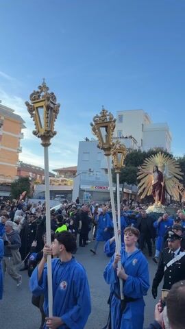 A religious procession is occurring in a European city with a crowd of people observing a statue of Jesus Christ being carried on a platform by men in blue robes. Two men in matching robes walk in front of the procession carrying tall, ornate golden lanterns on white poles. People on the sides of the road are watching, some with phones to capture the moment. Police officers and security personnel are present to manage the crowd. The sky is a clear blue with a few scattered clouds. Buildings of varying heights and architectural styles line the street. A grocery store sign is visible in the background.