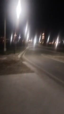 The video captures a dimly lit, empty asphalt road at night. Streetlights illuminate the scene, creating streaks of light and casting shadows. The road appears to curve to the right, with a sidewalk or path on the left. In the distance, several buildings with illuminated windows are visible, suggesting an urban or suburban setting. The overall atmosphere is quiet and deserted.
